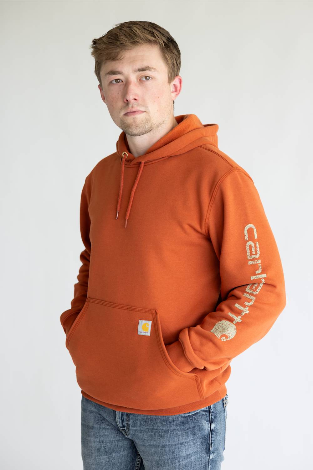 Man wearing a burnt orange colored hoodie, with Carhartt logo down the left arm sleeve. 