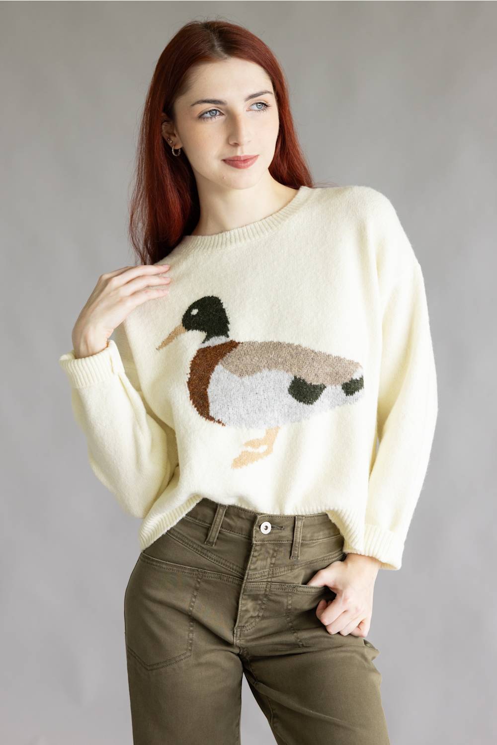 A woman with red hair wearing a cream-colored knit sweater with a detailed duck motif on the front, paired with olive green pants, standing against a neutral gray studio background.