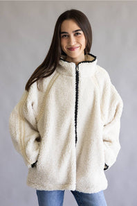A woman wearing a cream-colored sherpa fleece zip-up jacket with black trim along the zipper and collar, paired with blue jeans, set against a plain gray studio background.