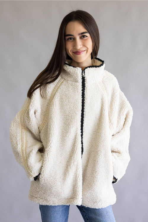 A woman wearing a cream-colored sherpa fleece zip-up jacket with black trim along the zipper and collar, paired with blue jeans, set against a plain gray studio background.