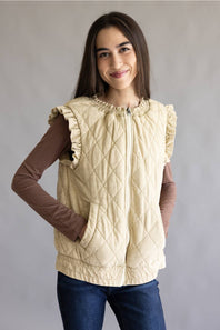 A young girl wearing a cream-colored quilted sleeveless vest with ruffle trim over a brown long-sleeve shirt and blue jeans, standing against a neutral gray studio background.