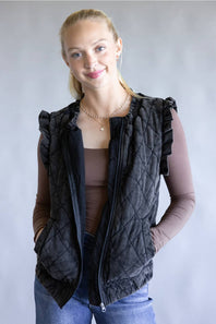 A woman modeling a black quilted sleeveless vest with ruffle trim over a brown long-sleeve top and blue jeans, standing against a neutral gray background.