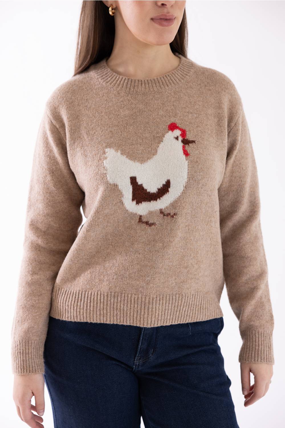 A woman wearing a beige crewneck sweater with a knitted white chicken graphic featuring a red comb and brown details, paired with dark blue jeans.