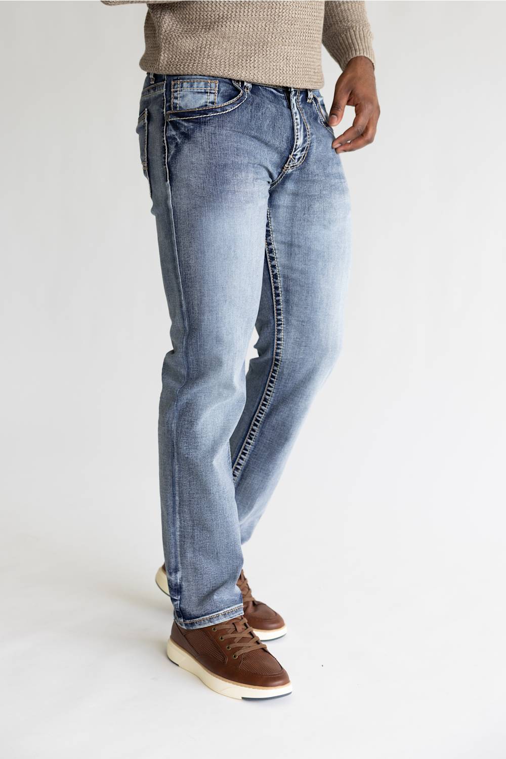 A person wearing light blue denim jeans with a faded wash, styled with a beige knit sweater and brown suede sneakers against a plain white studio background.