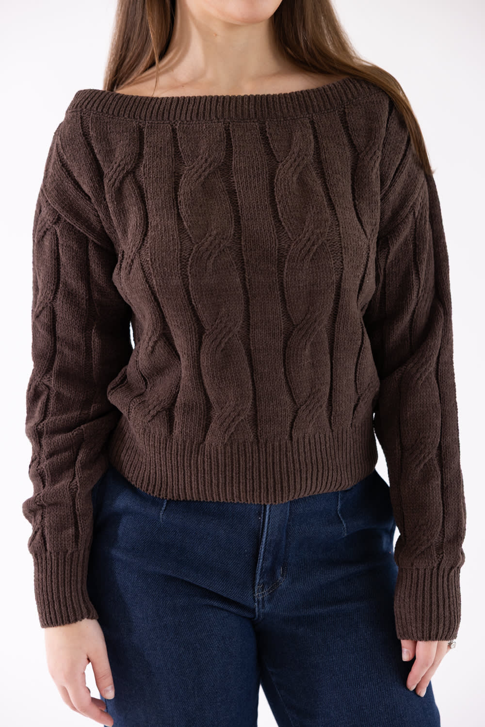 A woman wearing a dark brown cable-knit off-the-shoulder sweater and dark blue jeans, shown from the neck down against a plain white background.
