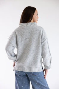 A woman shown from the back wearing a light gray knit sweater and blue jeans against a plain white studio background.