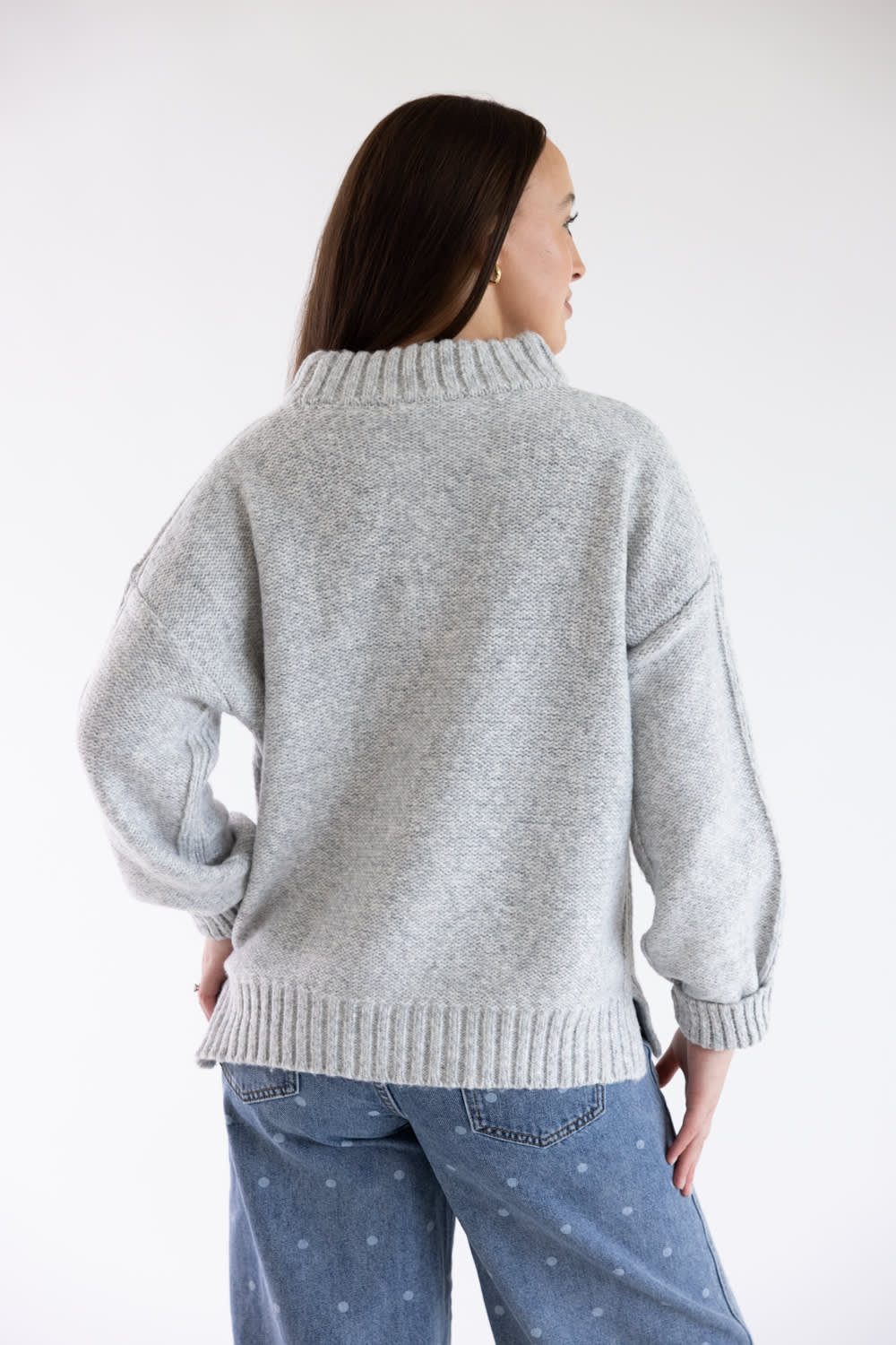 A woman shown from the back wearing a light gray knit sweater and blue jeans against a plain white studio background.