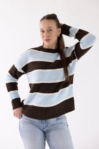 A woman modeling a brown, light blue, and white striped knit sweater with light-wash blue jeans, posing against a plain white studio background.