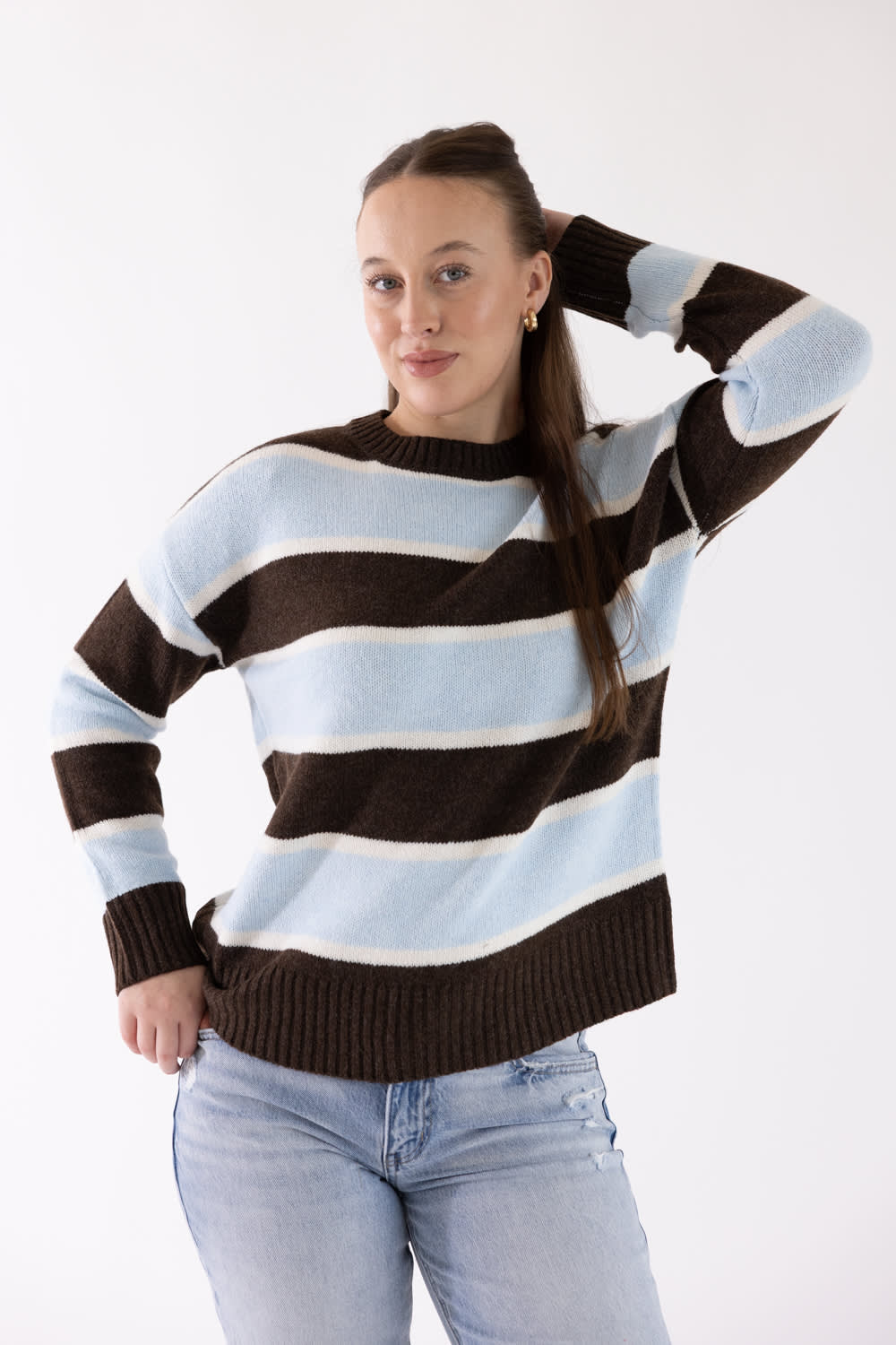 A woman modeling a brown, light blue, and white striped knit sweater with light-wash blue jeans, posing against a plain white studio background.