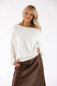 A woman modeling a fuzzy off-white knit sweater with a wide boat neckline, paired with a brown satin skirt, against a plain white studio background.