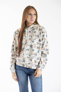 A woman with long light brown hair wearing a beige, brown, and gray camouflage patterned hoodie and blue jeans, standing against a plain white studio background.