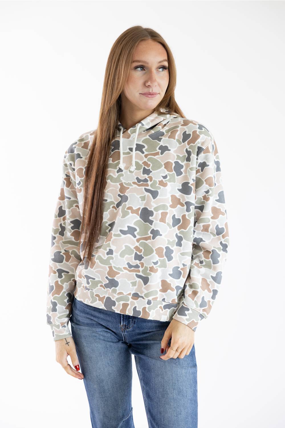 A woman with long light brown hair wearing a beige, brown, and gray camouflage patterned hoodie and blue jeans, standing against a plain white studio background.