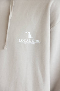 Close-up of a light beige hoodie with a white printed logo on the chest that reads 'LOCAL GIRL OUTFITTERS' with a small dog silhouette.