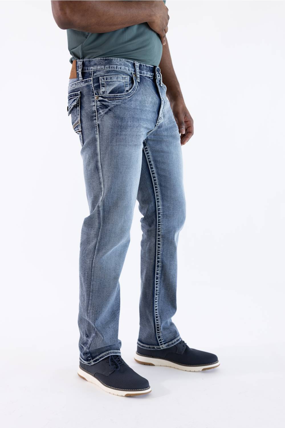 A person wearing light blue denim jeans with a faded wash, paired with a dark green shirt and navy shoes, shown from the waist down against a plain white studio background.