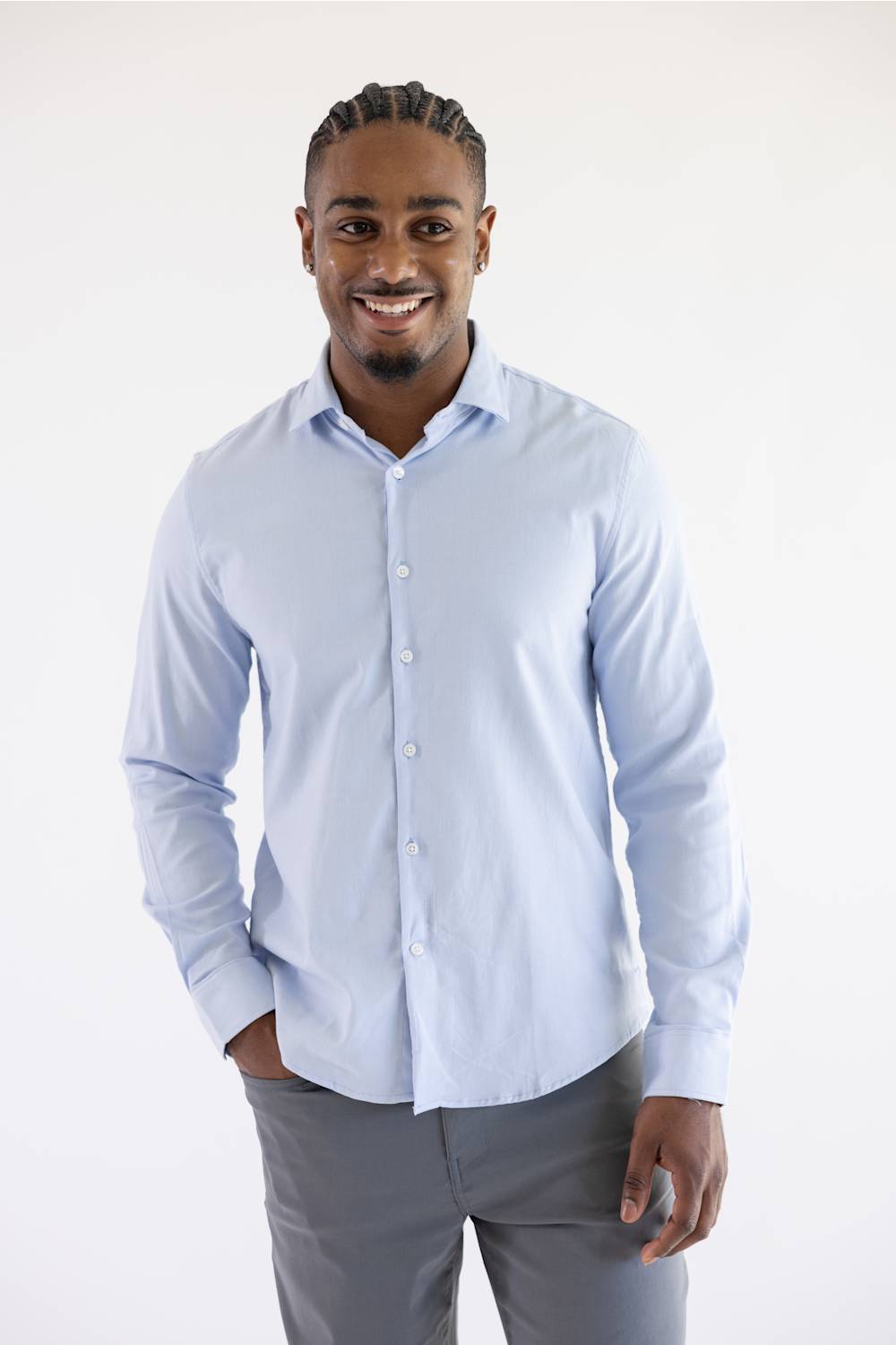 A smiling man wearing a light blue long-sleeve button-down shirt and gray trousers, standing against a plain white studio background.