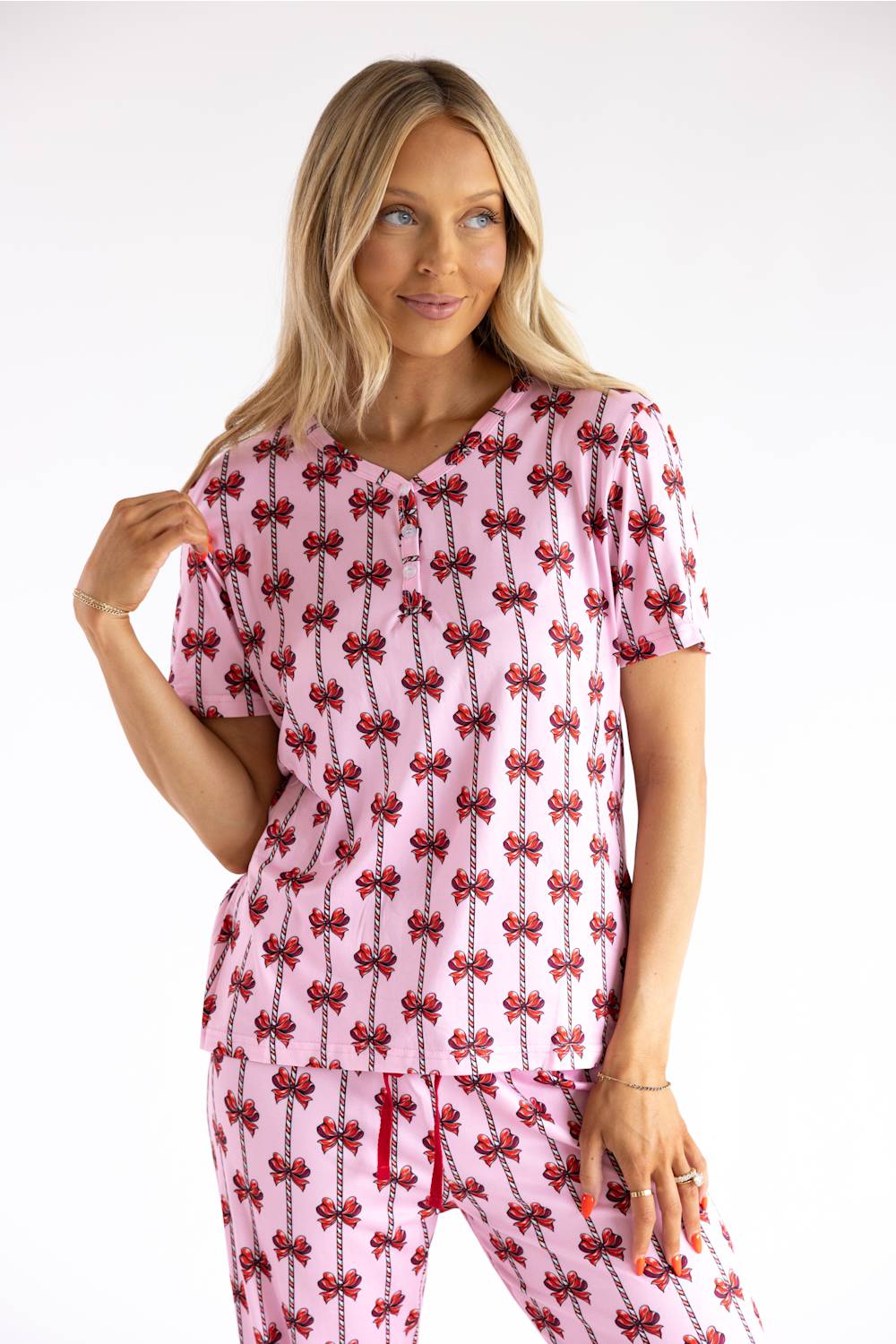 A woman modeling a pink short-sleeve scrub top and matching scrub pants set with a repeating red bow pattern, against a plain white studio background.