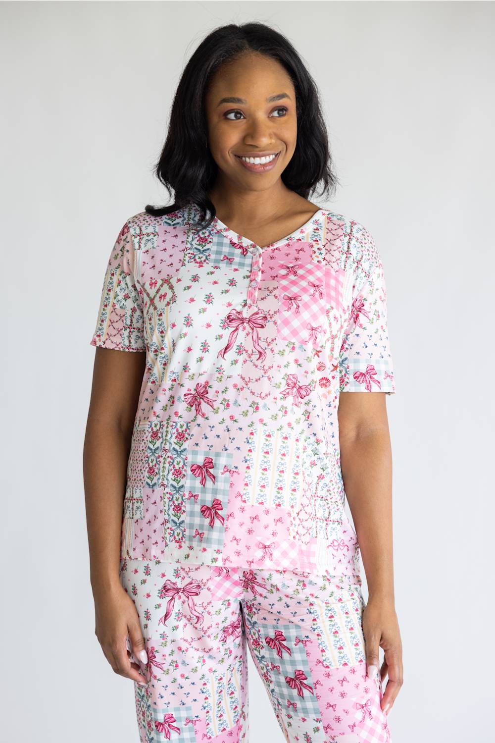 A woman modeling a matching two-piece pajama set with a patchwork print of pink bows, gingham, and floral motifs on a white background, photographed against a light gray studio backdrop.