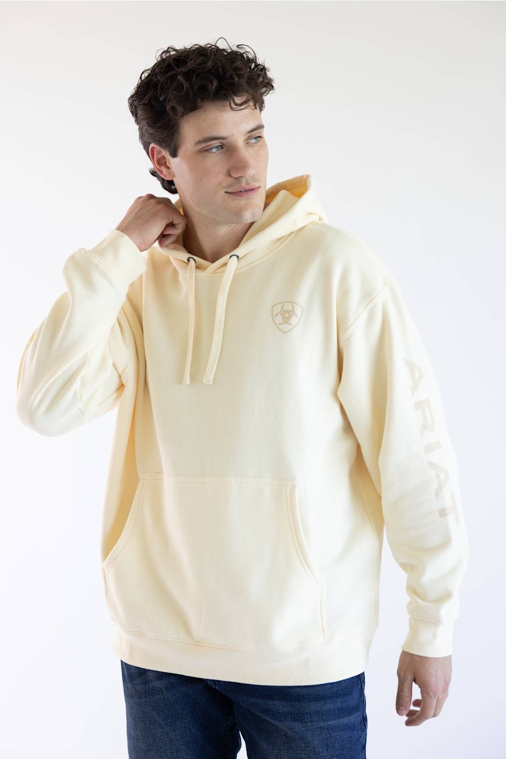 A man wearing a cream-colored hoodie with a small shield logo on the left chest 