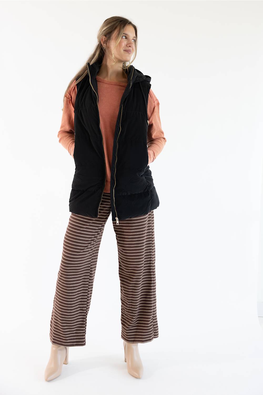 A woman modeling a black zip-up vest over a terracotta top and brown-and-brown-striped wide-leg pants, photographed against a clean white studio background.