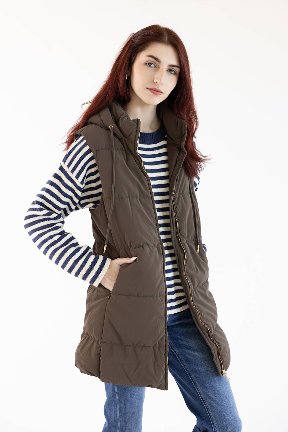 A woman with red hair wearing a brown hooded puffer vest over a navy and white striped long-sleeve shirt, paired with blue jeans, photographed against a plain white studio background.
