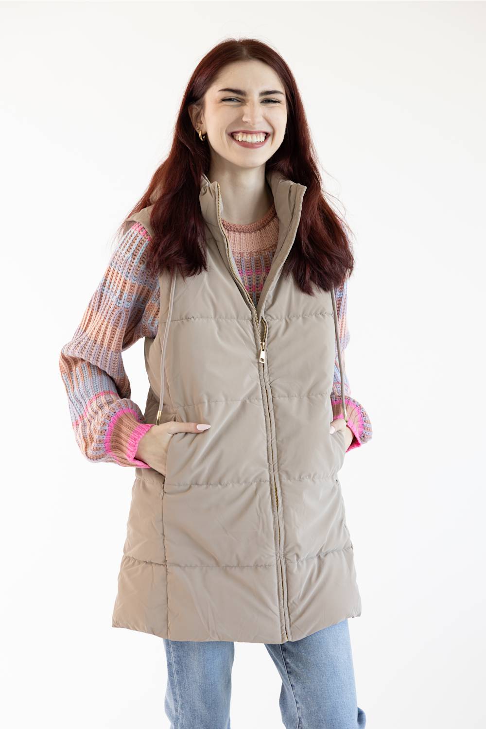 A woman smiling while wearing a beige sleeveless puffer vest over a pink and orange striped knit sweater, paired with light blue jeans, against a plain white background.