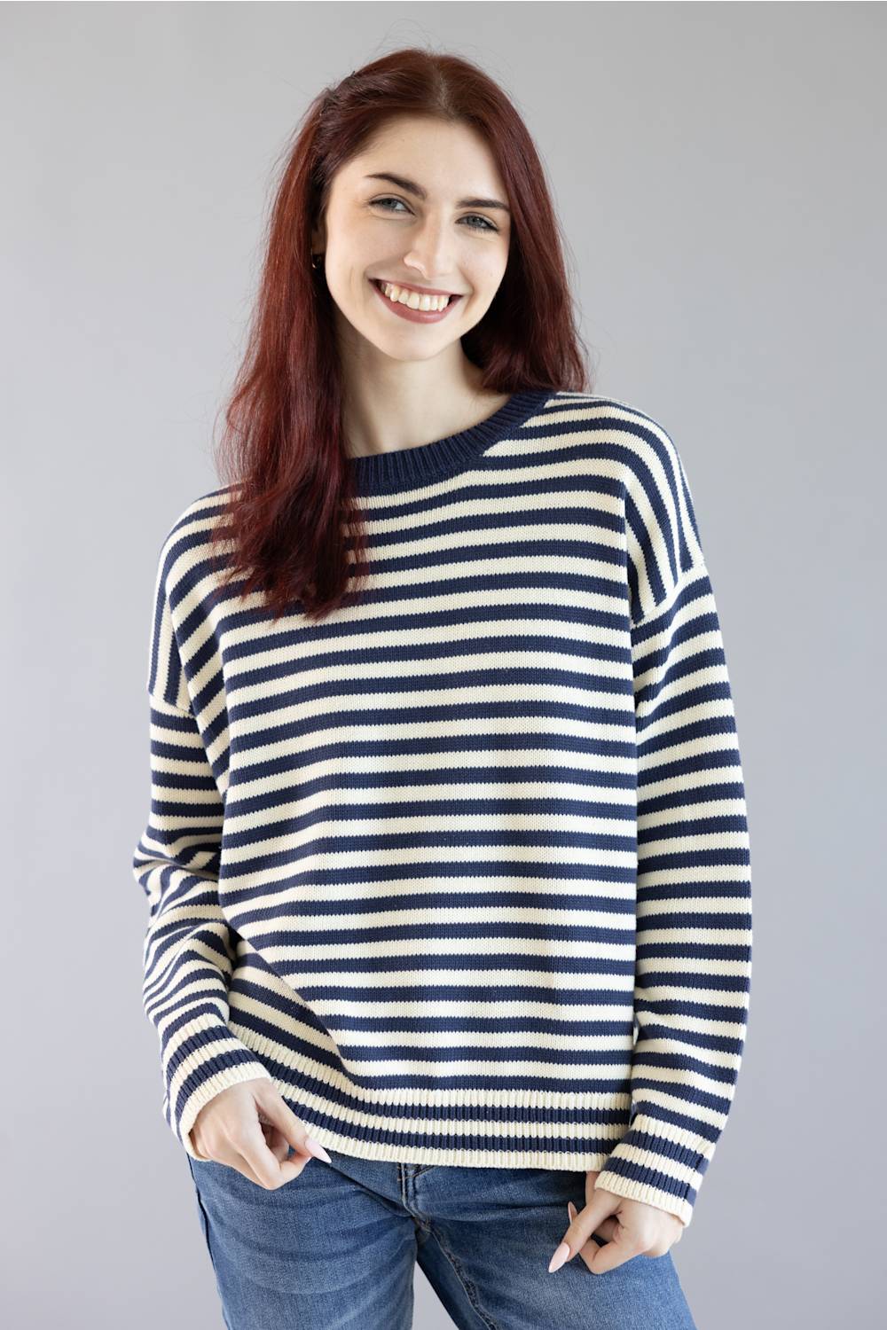 A smiling woman with red hair wearing a navy and cream horizontally striped knit sweater and blue jeans, standing against a solid gray studio background.