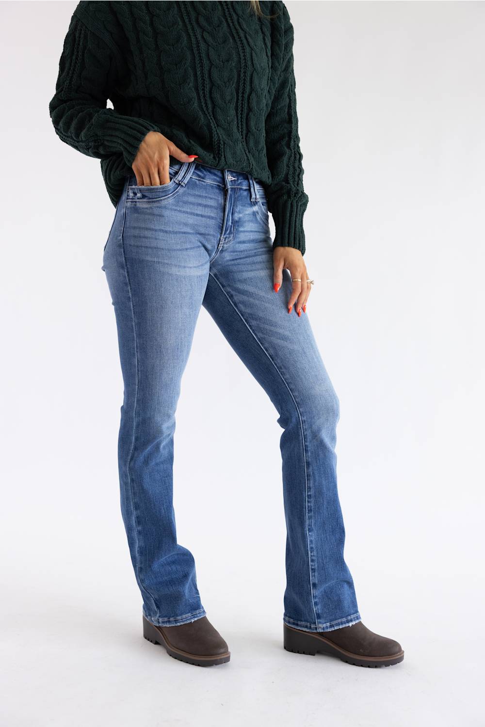 A model wearing blue flare jeans, a dark green cable-knit sweater, and brown boots, standing against a plain white studio background.