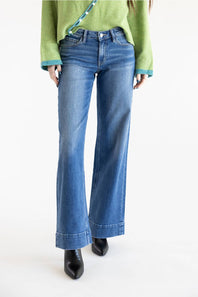 A person wearing medium-wash blue flare jeans with a green sweater and black pointed shoes, photographed against a clean white studio background.