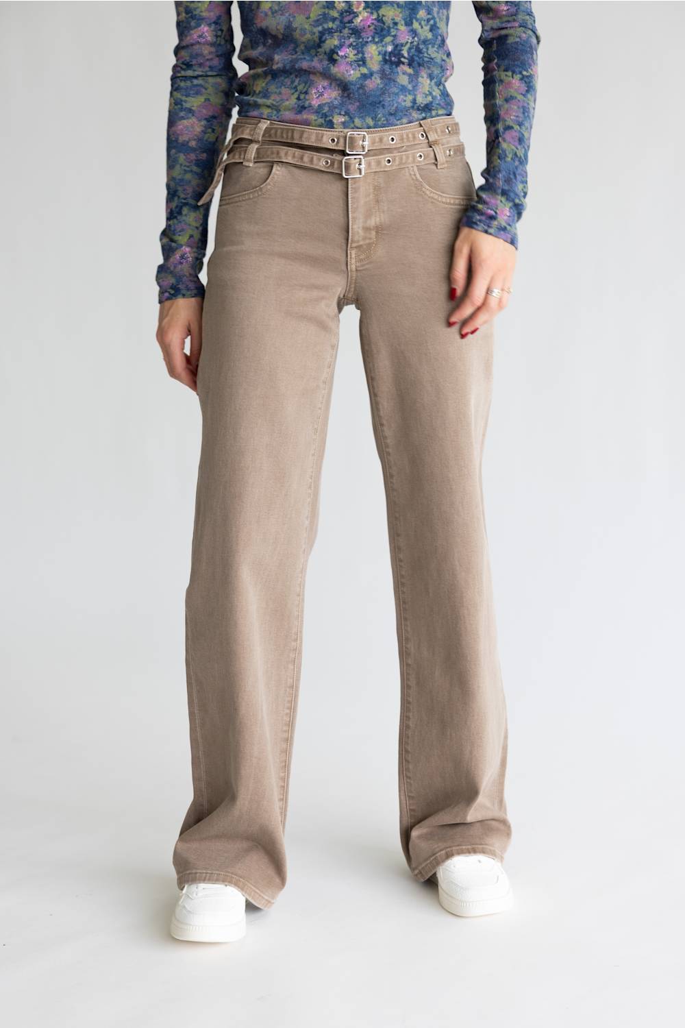 A model is wearing a pair of wide-leg beige jeans with a matching belt, styled with a floral long-sleeve shirt and white sneakers against a plain light gray background.