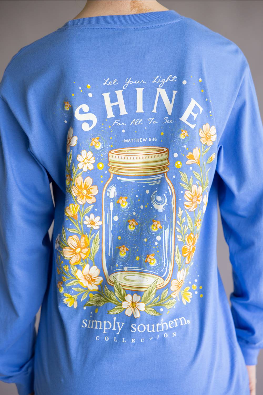 A person wearing a blue long-sleeve shirt, shown from the back. The shirt features a large graphic print of a mason jar surrounded by yellow and white flowers, with the text 'Let Your Light SHINE' and 'simply southern COLLECTION'.