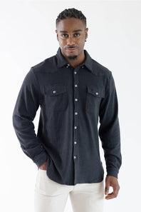 A man wearing a dark charcoal gray long-sleeve western-style button-up shirt with two chest pockets, paired with white pants, photographed against a plain white studio background.