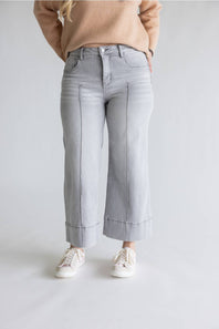 A person wearing light gray wide-leg cropped jeans with rolled cuffs, a beige sweater, and white sneakers, standing against a light gray studio background.