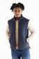 A man wearing a navy blue diamond-quilted sleeveless puffer vest over a beige crewneck sweater and blue jeans, standing against a plain white background.