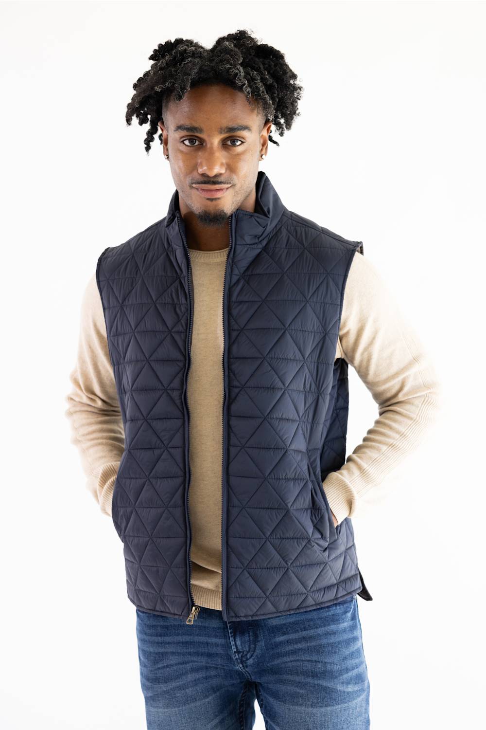 A man wearing a navy blue diamond-quilted sleeveless puffer vest over a beige crewneck sweater and blue jeans, standing against a plain white background.