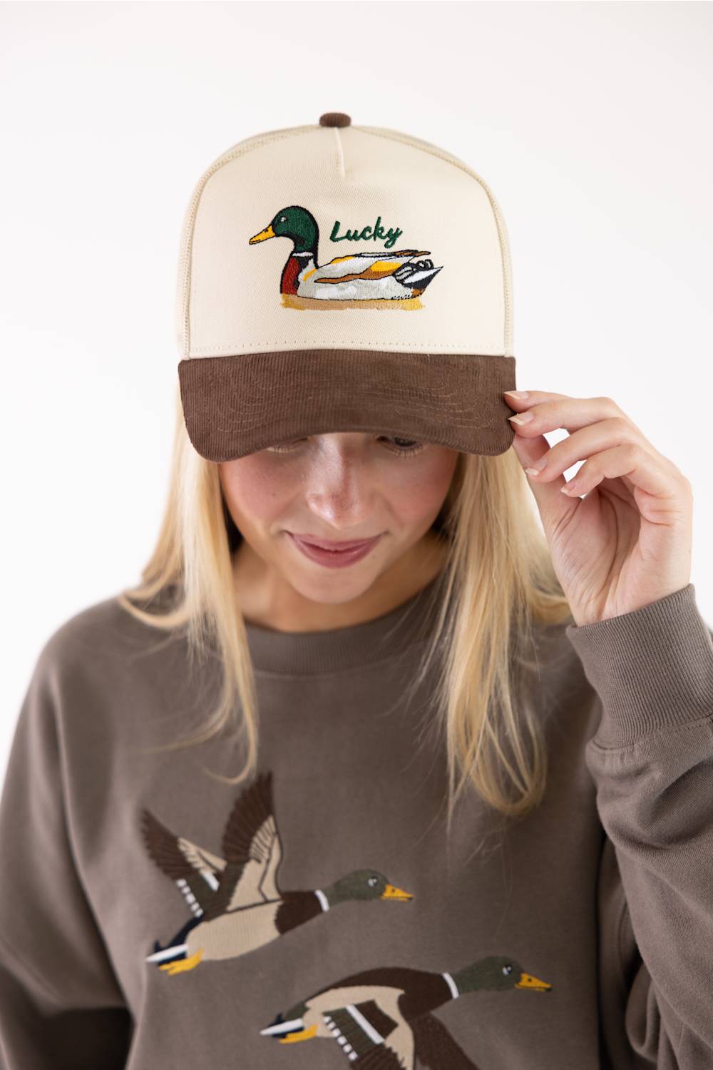 A woman wearing a cream and brown trucker hat with an embroidered mallard duck and the word 'Lucky' on the front, paired with a brown sweatshirt featuring mallard graphics, against a plain white background.