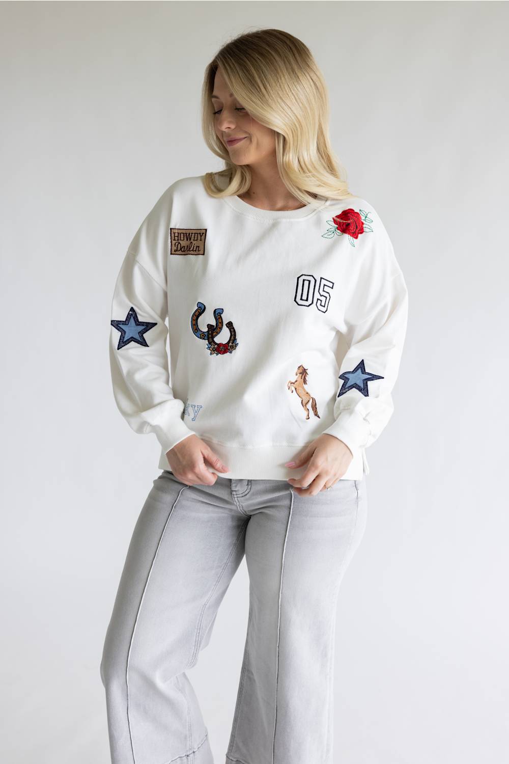 A woman wearing a white crewneck sweatshirt with various embroidered patches, including a red rose, blue stars, a horseshoe, and the number '05', paired with light gray jeans, against a plain light gray background.