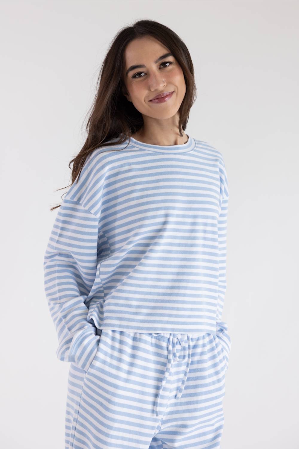 A woman wearing a light blue and white striped long-sleeve top and matching striped pants, standing against a plain light gray studio background.
