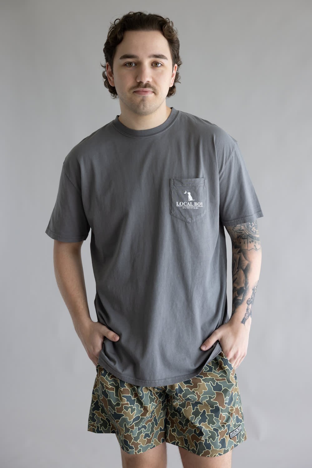Local Boy Vintage Emblem T-Shirt for Men in Grey; Man wearing a gray t-shirt with a pocket and patterned shorts on a plain background