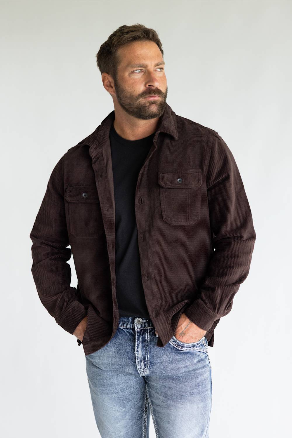 A man wearing a dark brown corduroy overshirt jacket over a black t-shirt and blue jeans, standing against a light gray studio background.