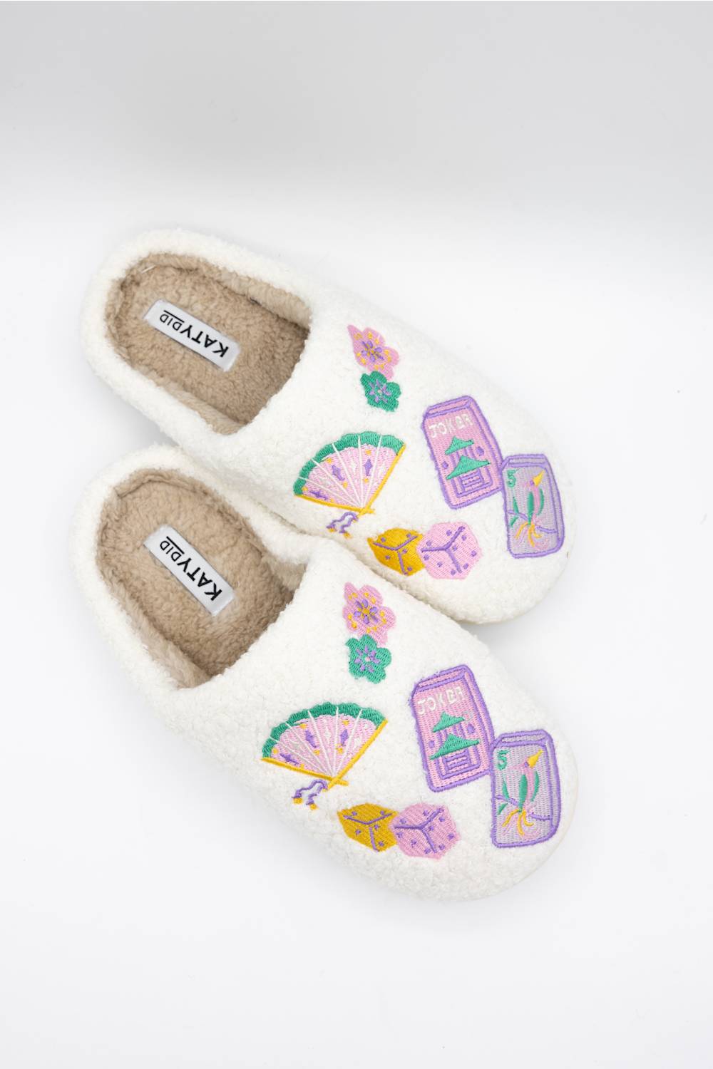 A pair of white sherpa slip-on slippers decorated with colorful embroidered majong and fan motifs on the sides, shown on a clean white background.