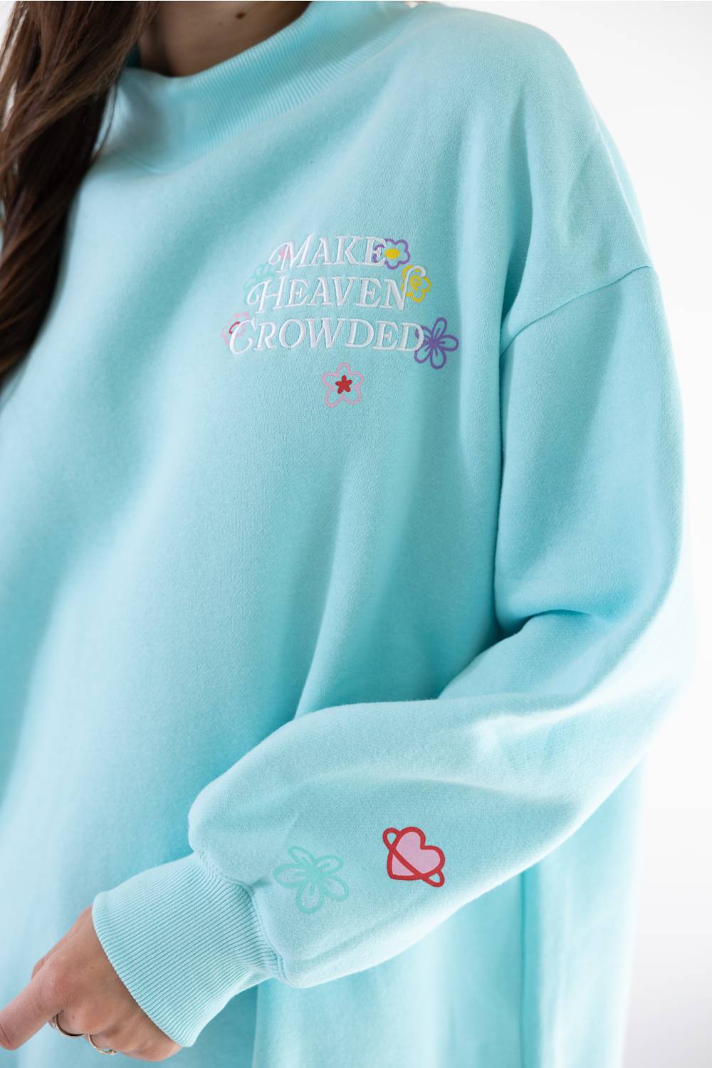 A close-up of a person wearing a light blue sweatshirt with colorful embroidered text reading 'MAKE HEAVEN CROWDED' on the chest and small floral/heart embroidery on the sleeve.