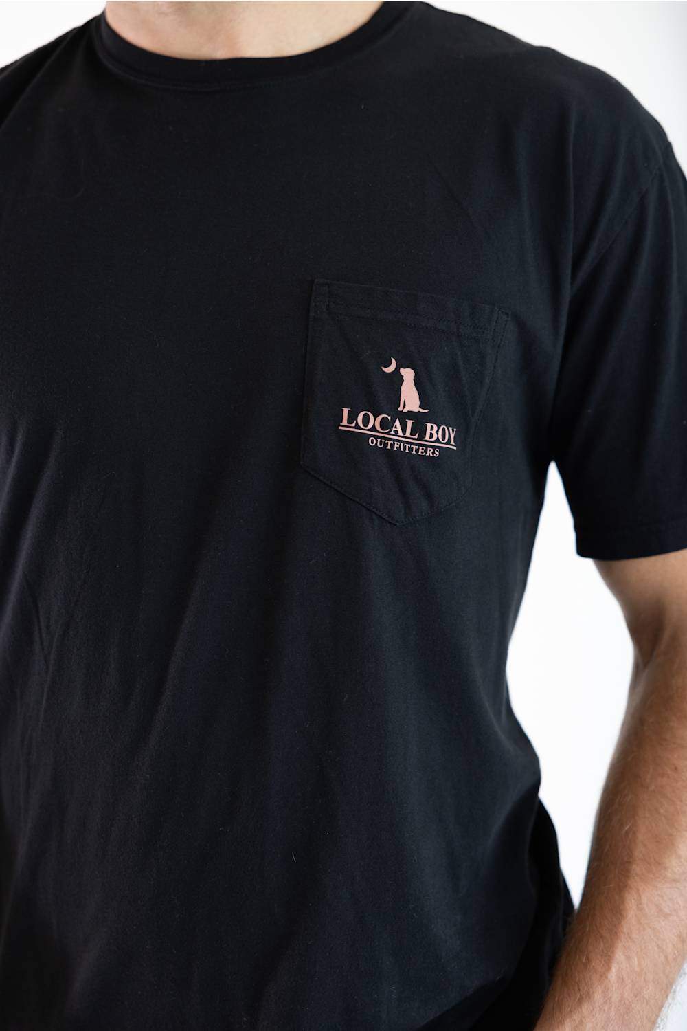 A close-up of a person wearing a black short-sleeve pocket t-shirt with a chest pocket featuring a pink and gold 'LOCAL BOY ' graphic.