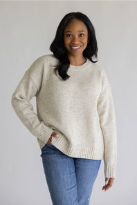 A woman wearing a cream-colored, speckled knit crewneck sweater and blue jeans, standing against a plain light gray background.