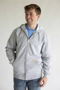 A man wearing a light gray zip-up hoodie with a drawstring hood and a small yellow Carhartt logo patch on the pocket, layered over a light blue t-shirt and blue jeans, standing against a plain light gray wall.