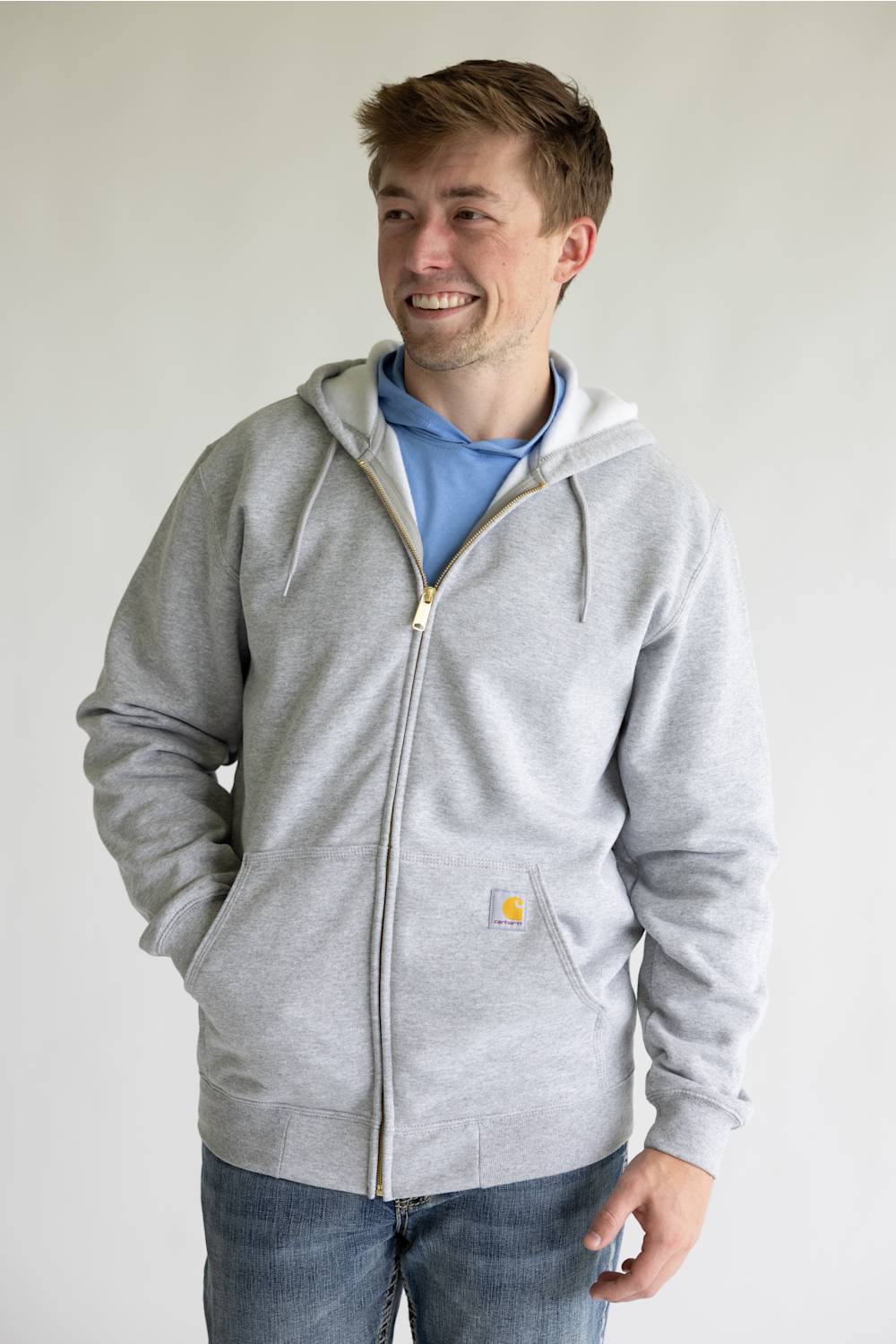 A man wearing a light gray zip-up hoodie with a drawstring hood and a small yellow Carhartt logo patch on the pocket, layered over a light blue t-shirt and blue jeans, standing against a plain light gray wall.