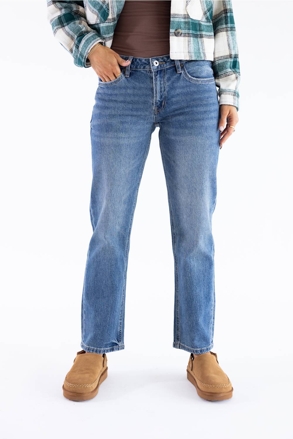 A person wearing medium-wash blue straight-leg jeans, styled with a brown top, a green and white plaid flannel shirt, and tan suede boots, shown from the waist down on a white studio background.