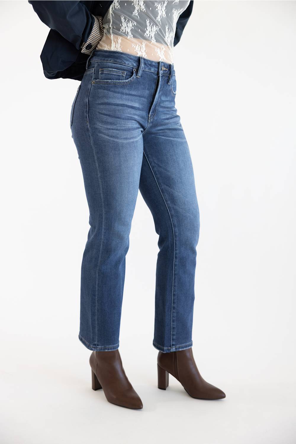 A model wearing a pair of dark-wash blue straight-leg jeans, styled with a white lace top, a dark navy jacket, and brown ankle boots, set against a plain white studio background.
