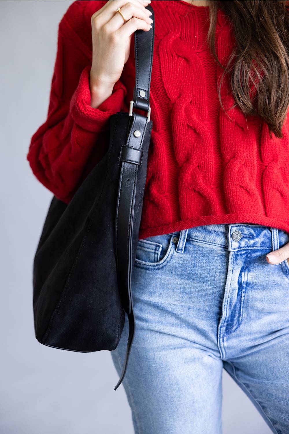 A person wearing a bright red cable-knit sweater and light blue jeans carries a large black shoulder bag with a black strap and silver-tone hardware against a plain light gray background.