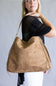 A woman wearing a black mesh top and white pants carries a large tan suede hobo shoulder bag against a plain gray studio background.