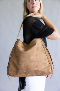 A woman wearing a black mesh top and white pants carries a large tan suede hobo shoulder bag against a plain gray studio background.